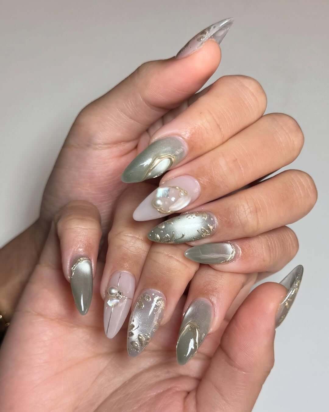 Cat eye almond-shaped nails with sage green swirls, milky neutral base, pearl accents, and gold foil details for an elegant earthy look.