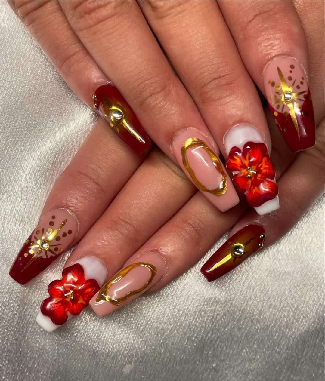 Long square nails with red 3D florals, gold foil accents, and gemstone embellishments.