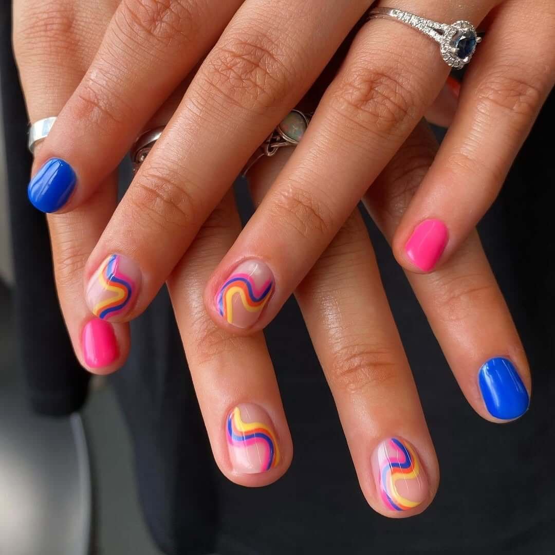 Short round nails with hot pink, electric blue, and rainbow wave patterns over a sheer base.